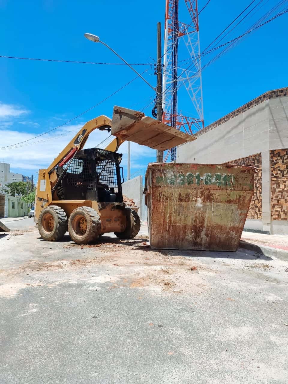 Minicarregadeira ao lado de caçamba de entulho na rua