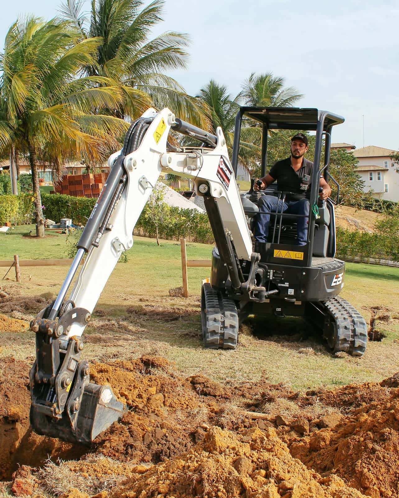 Mini escavadeira escavando num jardim de condomínio, com palmeiras ao fundo