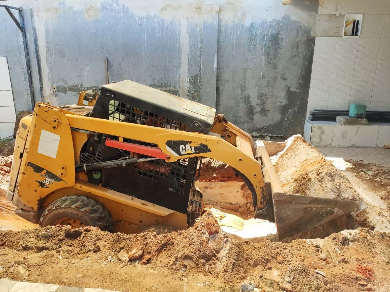 Minicarregadeira escavando em quintal cercado por muros